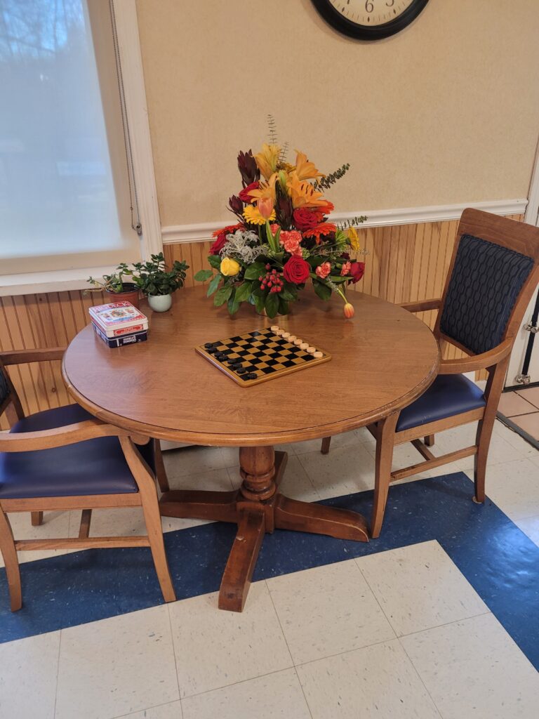 Round wooden game table with checkerboard, surrounded by two chairs and a vibrant floral arrangement, reflecting a welcoming atmosphere for seniors at Amelia Grace Assisted Living.
